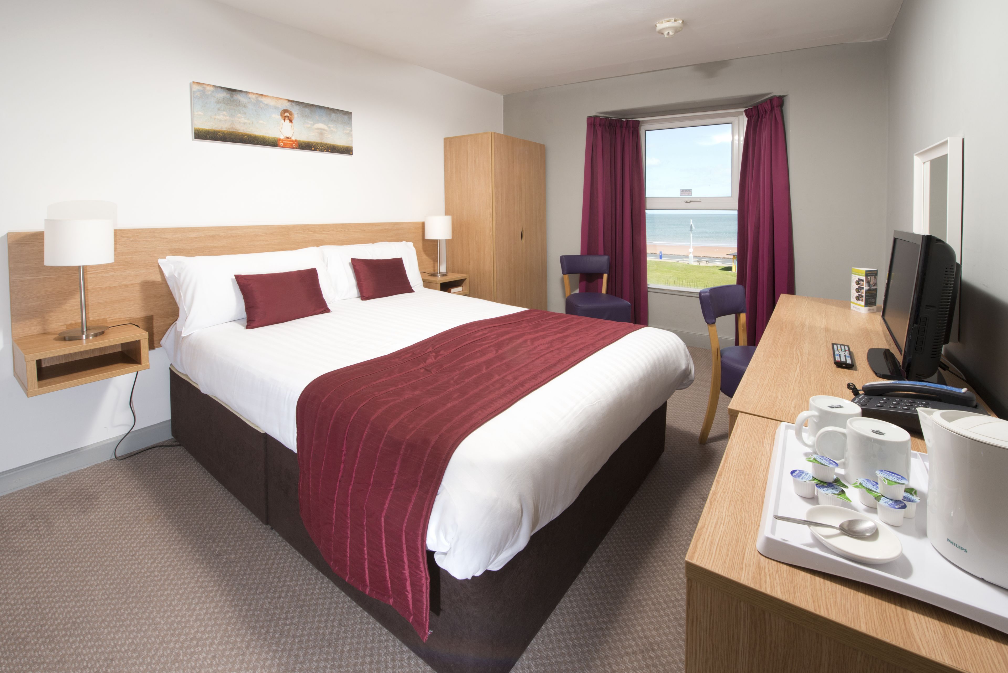 A hotel room with a large bed, a desk with a computer, a chair, and a window with a view of the beach.