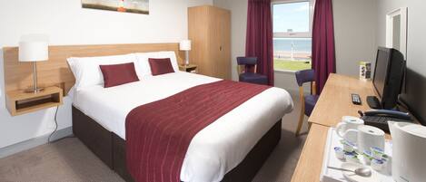 A hotel room with a large bed, a desk with a computer, a chair, and a window with a view of the beach.