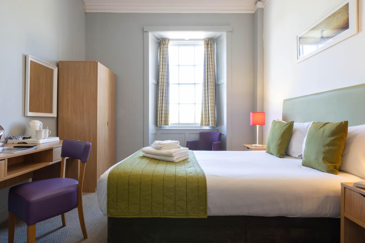 A hotel room with a bed, a desk, a chair, a window with curtains, and a nightstand with a lamp.