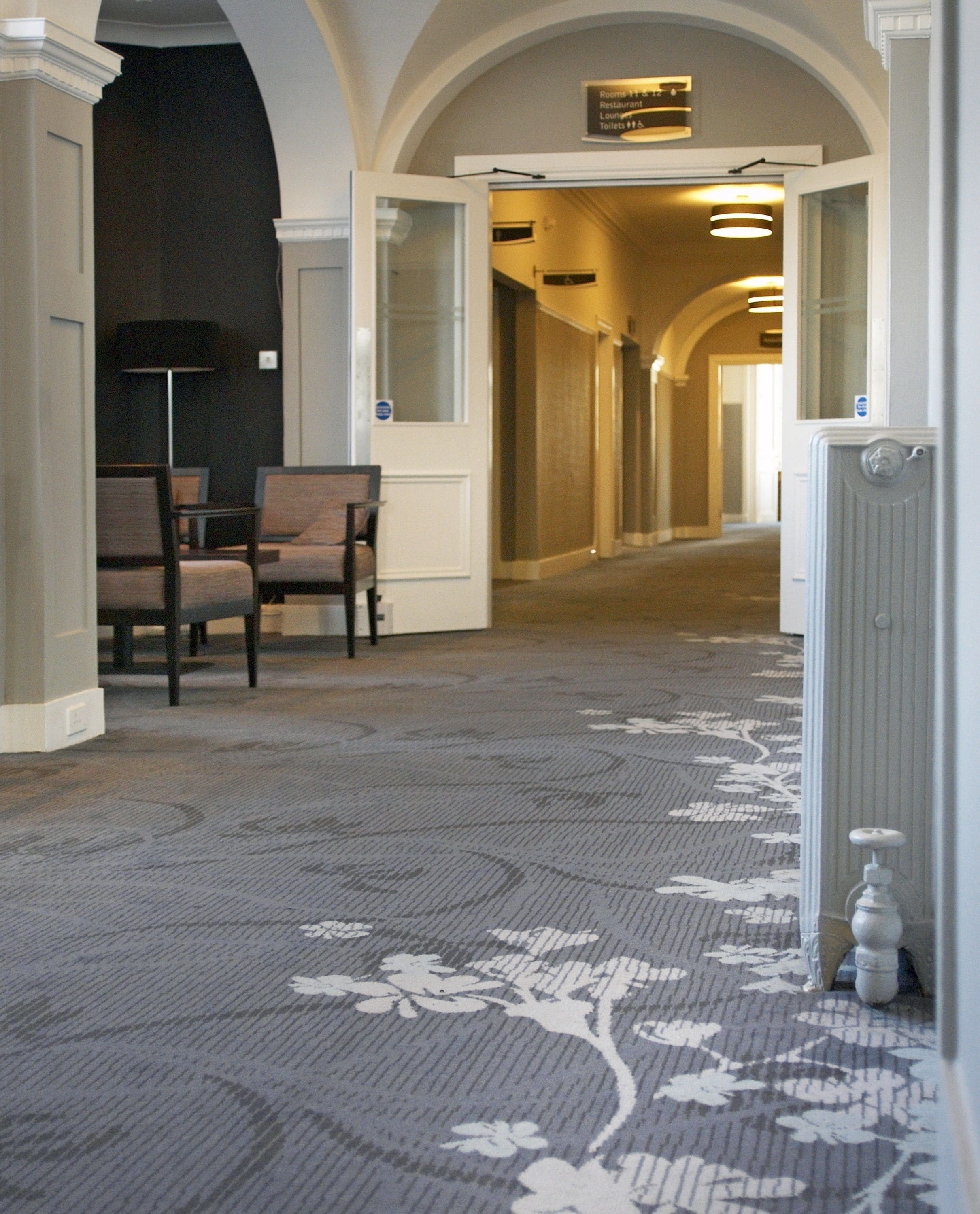 Pasillo de hotel con alfombra estampada, mostrador de recepción y señalización que indica la ubicación de habitaciones y baños.