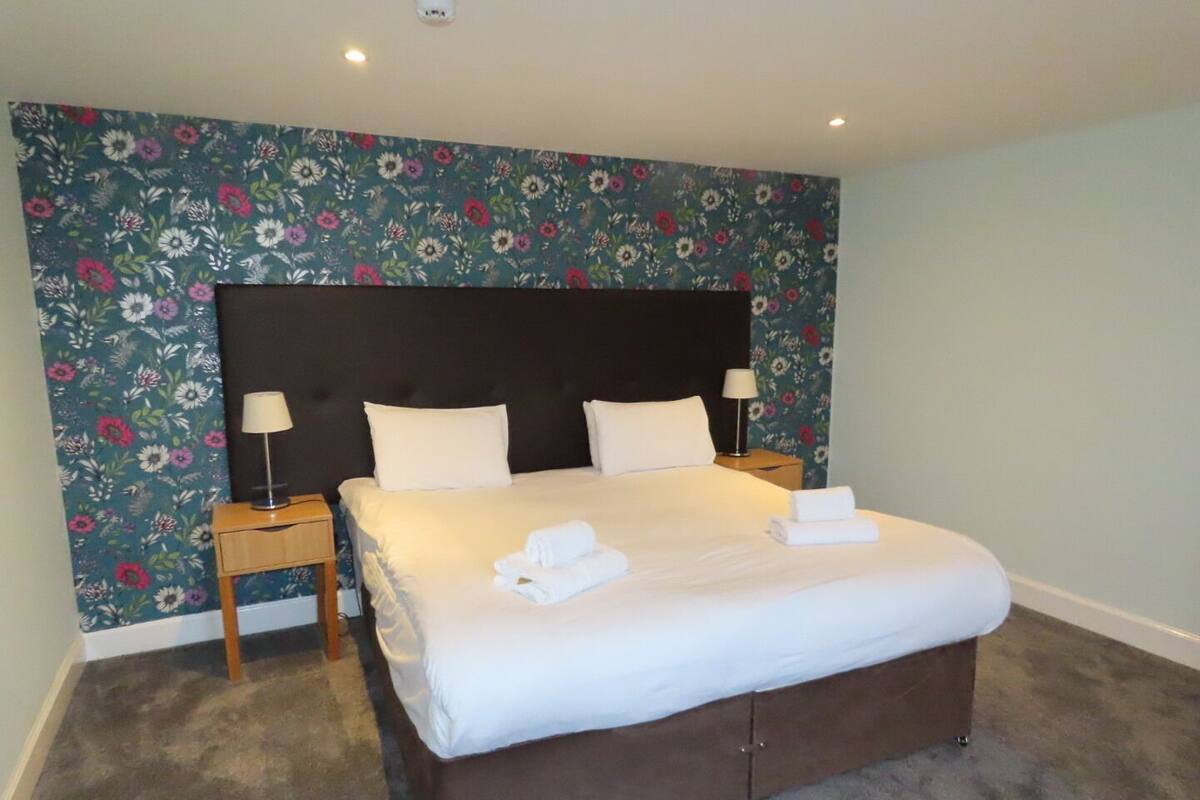 A neatly made bed with white linens and two pillows, a wooden nightstand with a lamp, and a floral wallpaper backdrop.