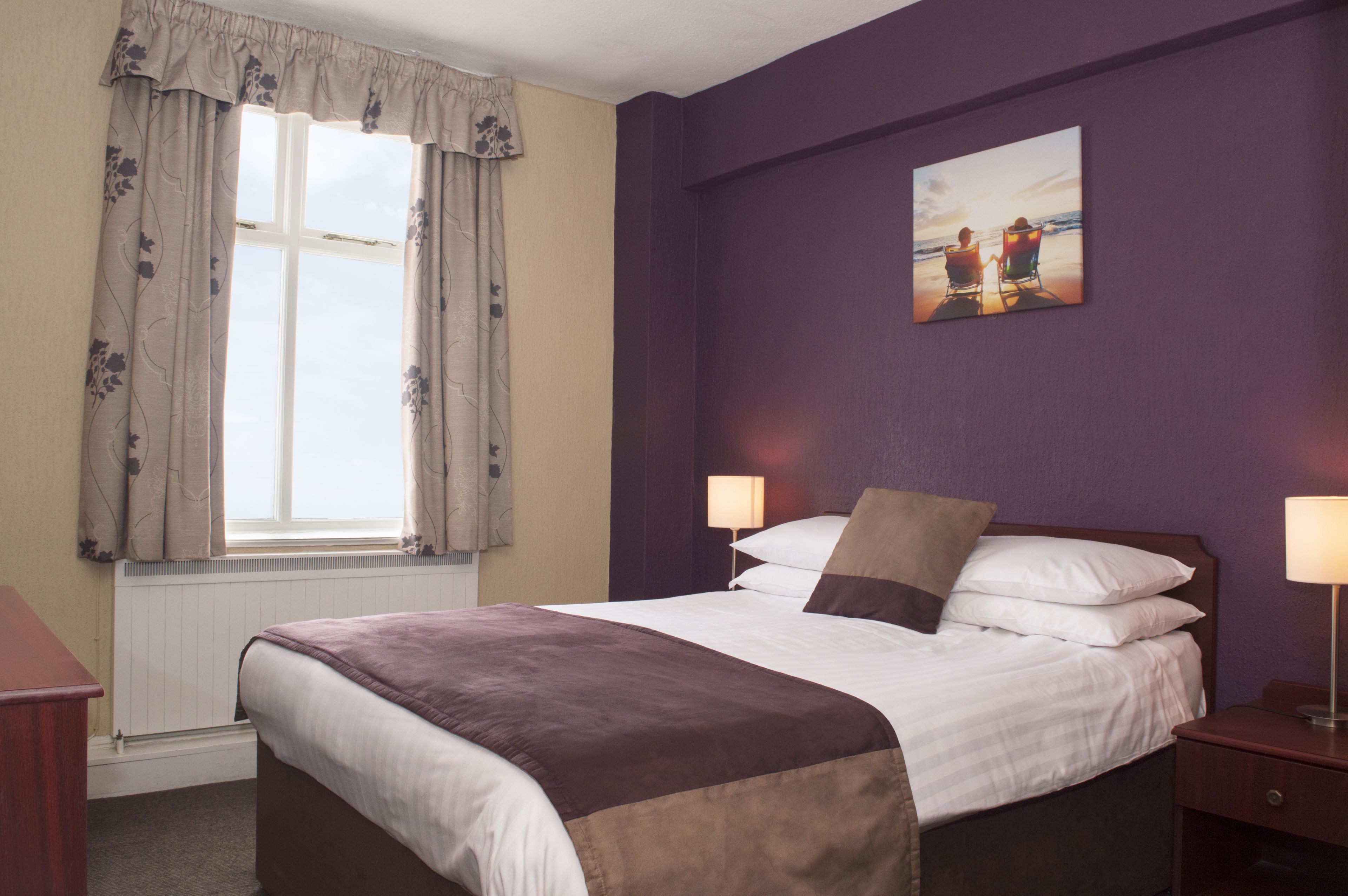 A hotel room with a large bed, two bedside lamps, a window with curtains, and a painting on the wall.
