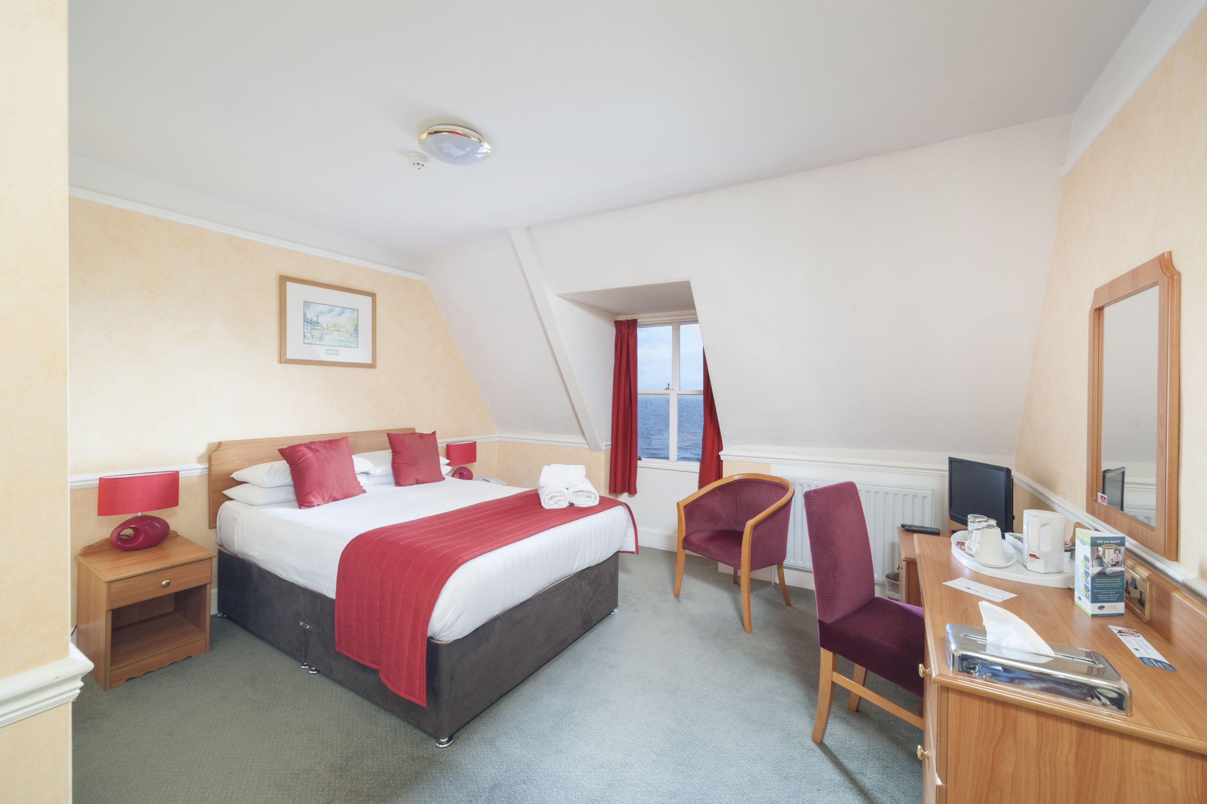 A hotel room with a bed, a desk, a chair, a nightstand, a lamp, a window, and a mirror.