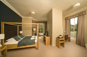 A four-poster bed with a wooden frame, a desk, and a chair in a room with large windows and a view of greenery outside.