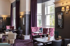 A restaurant with a purple carpet, tables with chairs, and a view of greenery outside. - Waverley Castle Hotel (Melrose)
