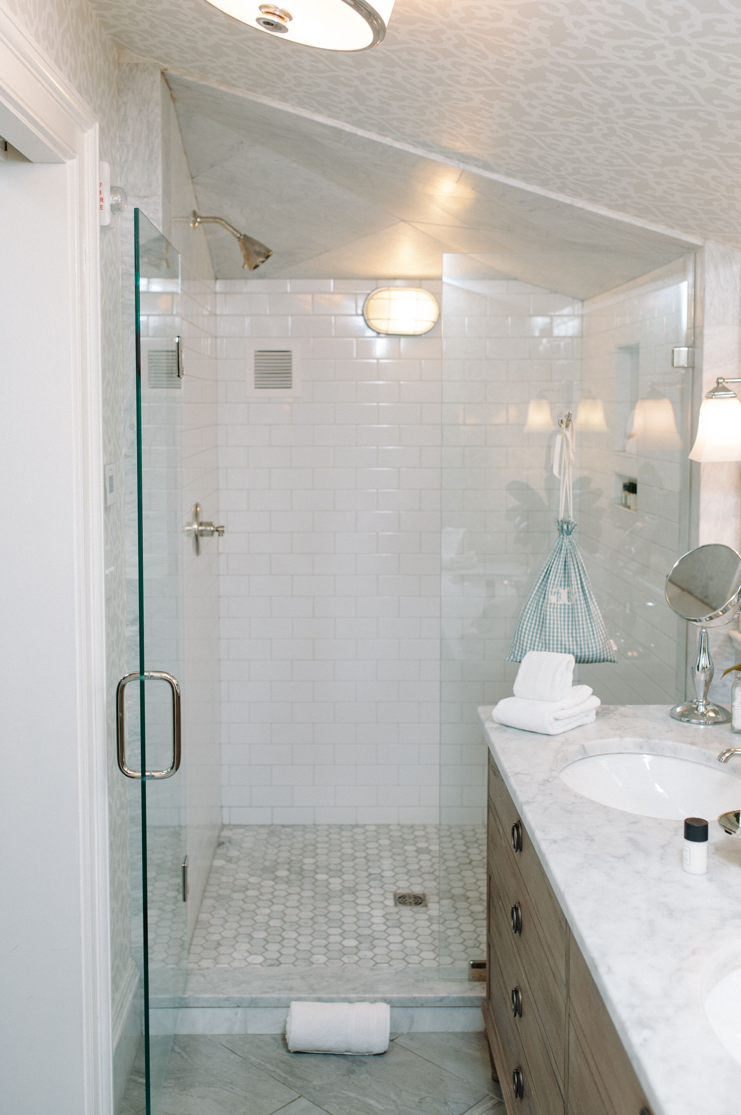 A modern bathroom with a glass-enclosed shower, white tiled walls, and a marble countertop with a sink and mirror.