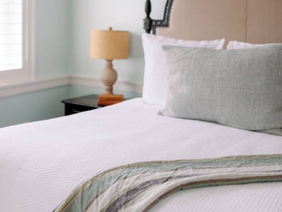 A neatly made bed with a headboard, a bedside lamp, and a window with blinds.