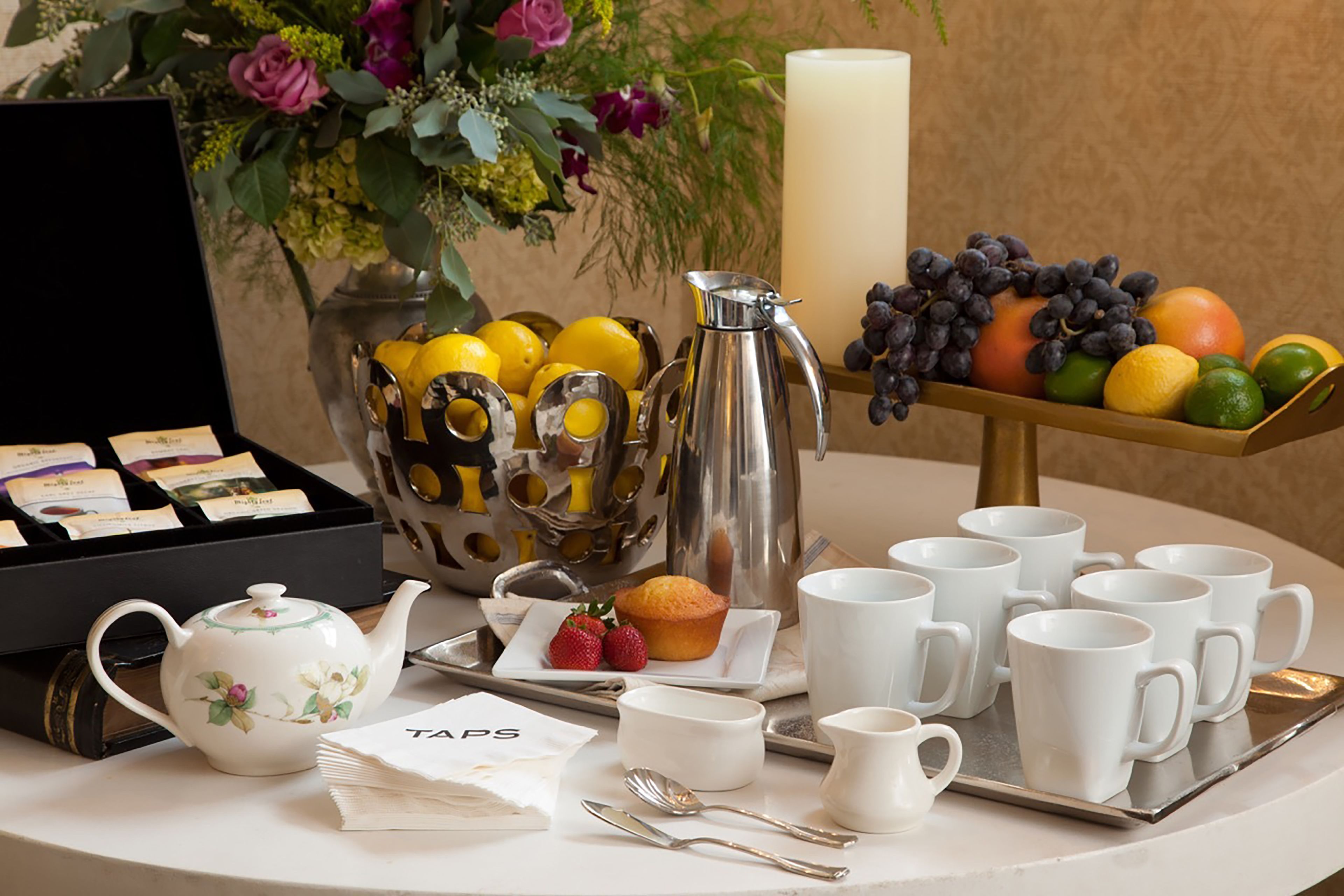 Uma mesa de café da manhã com chaleira, xícaras, bandeja com frutas, vela e um vaso de flores.