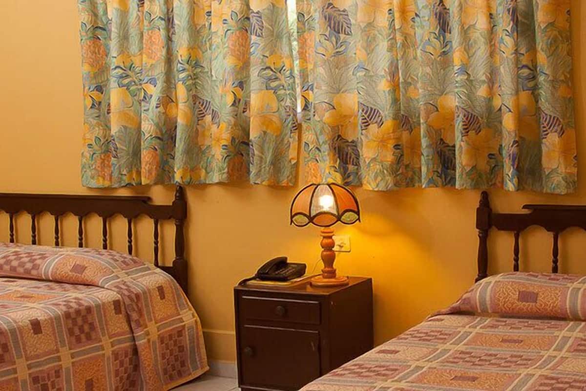 A hotel room with two beds, a nightstand with a lamp, and a window with floral curtains.