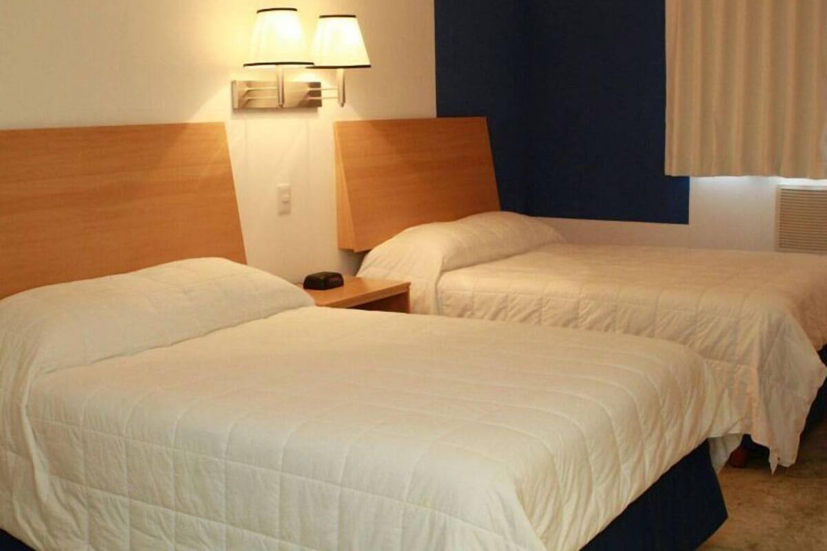 Habitación de hotel con dos camas, cabecero de madera, mesita de noche y lámpara de pared.