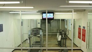 Uma academia com esteiras e equipamentos de ginástica, uma televisão na parede e uma escada que leva a um andar superior.