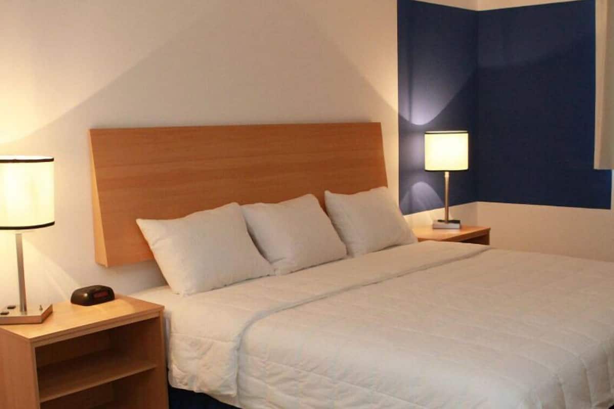 Una cama bien hecha con cabecera de madera, una mesita de noche con lámpara y un estante fijado a la pared.