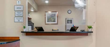 A hotel reception desk with a sign indicating doors are locked 24 hours, check-in at 3:00 PM, and check-out at 10:00 AM. There is a no smoking sign and a clock on the wall.