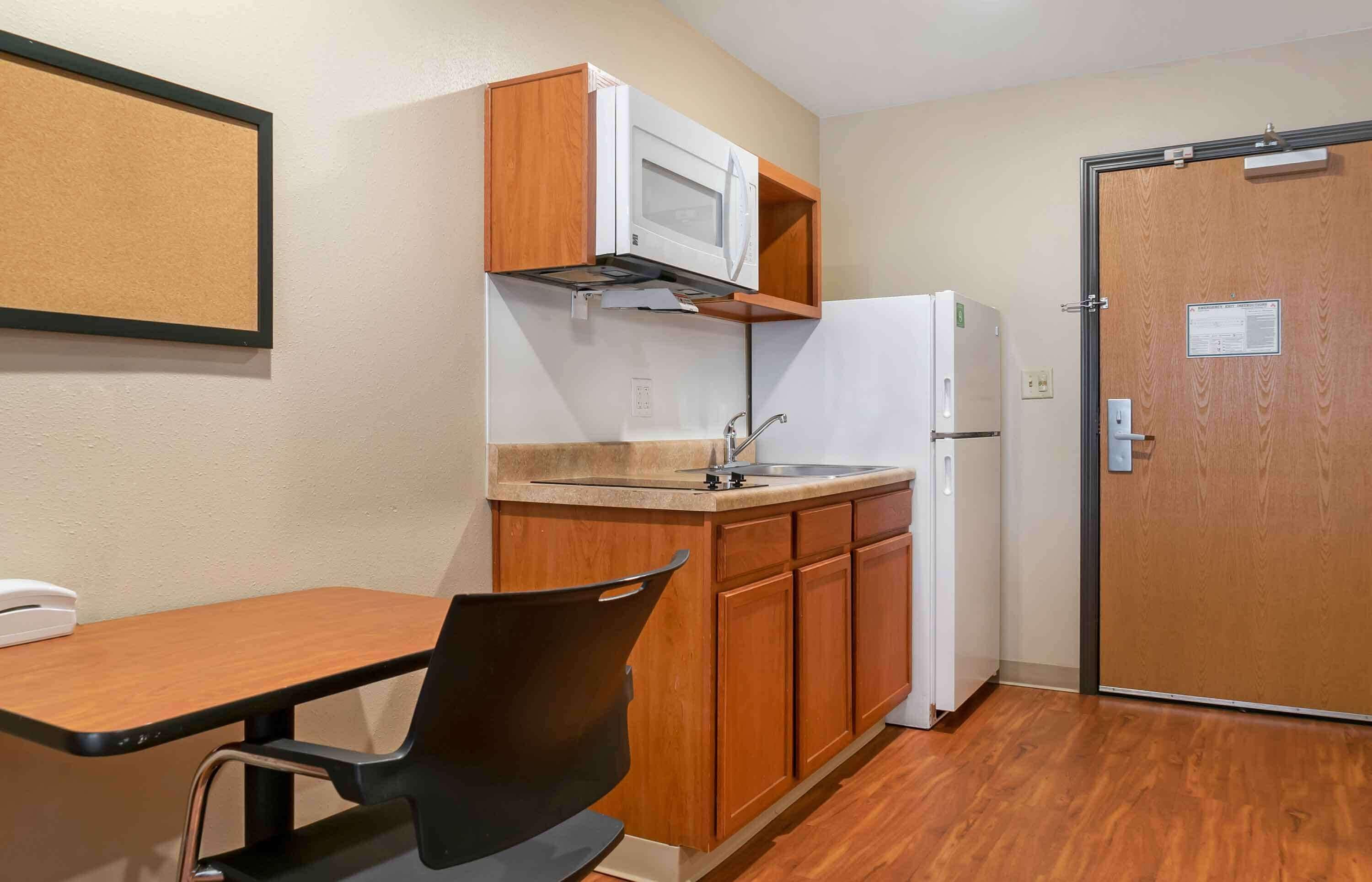 Una kitchenette compacta con microondas, pileta y refrigerador. Hay una mesa y silla de madera, y una puerta con un cartel.