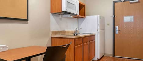 Una kitchenette compacta con microondas, pileta y refrigerador. Hay una mesa y silla de madera, y una puerta con un cartel.