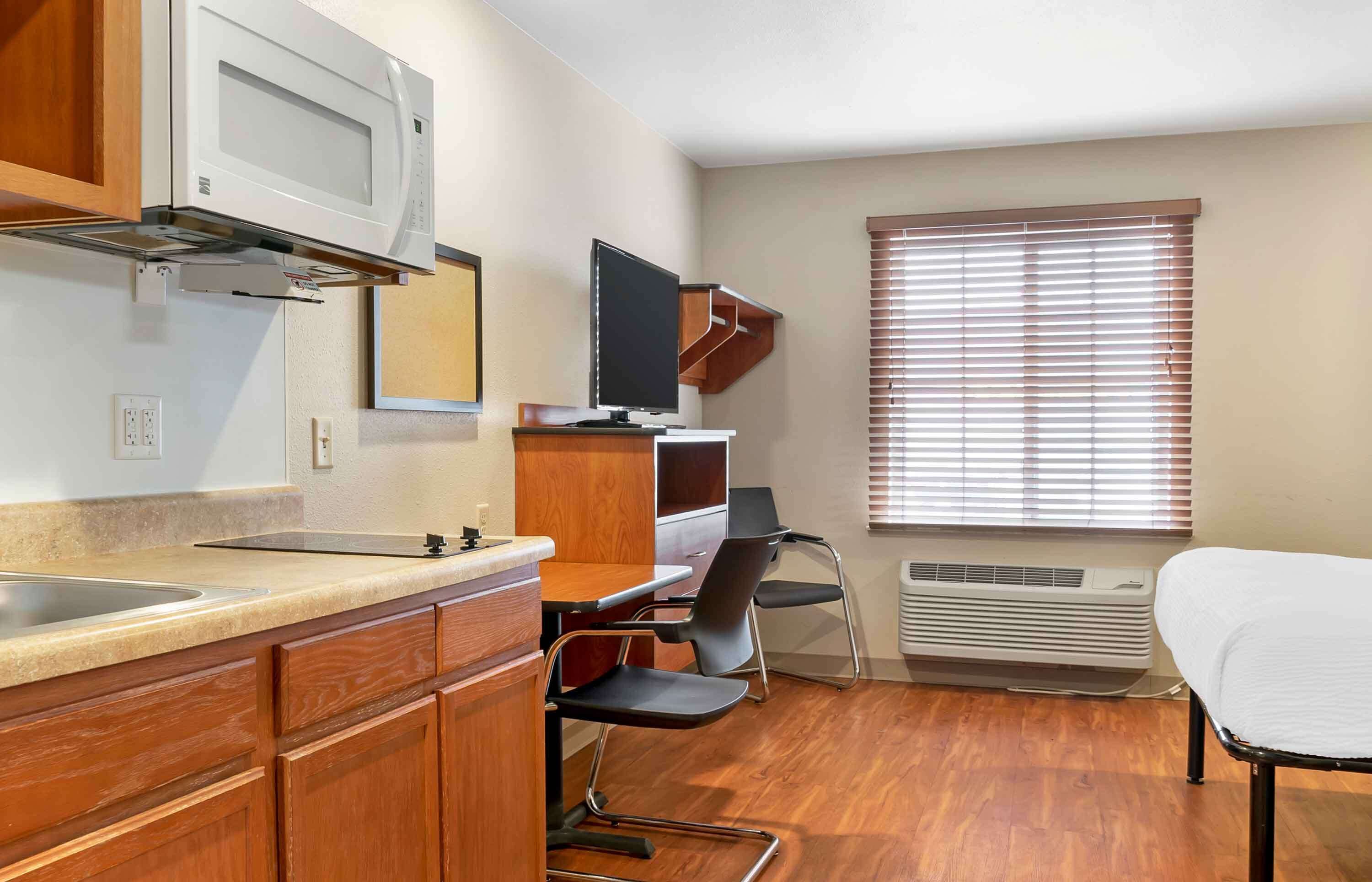 A compact kitchenette with a microwave, sink, and wooden cabinets. There is a TV mounted on the wall and a window with blinds.