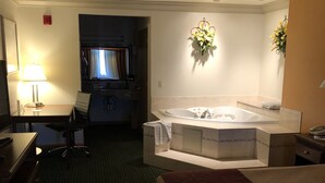 Habitación de hotel con cama, escritorio, silla, mesita, lámpara, televisor y baño con bañera. También hay un arreglo floral en la pared.