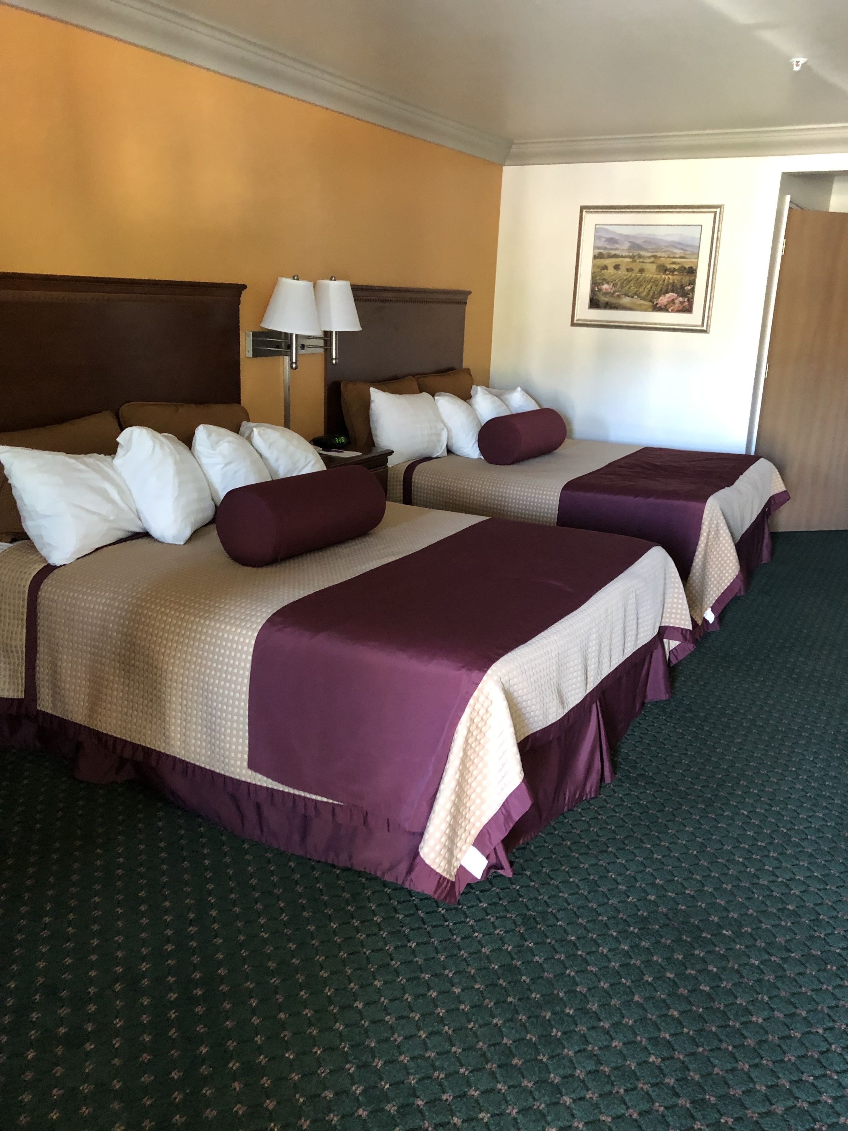 Une chambre d’hôtel avec deux lits, une table de chevet avec une lampe, un tableau au mur et un sol recouvert de moquette.