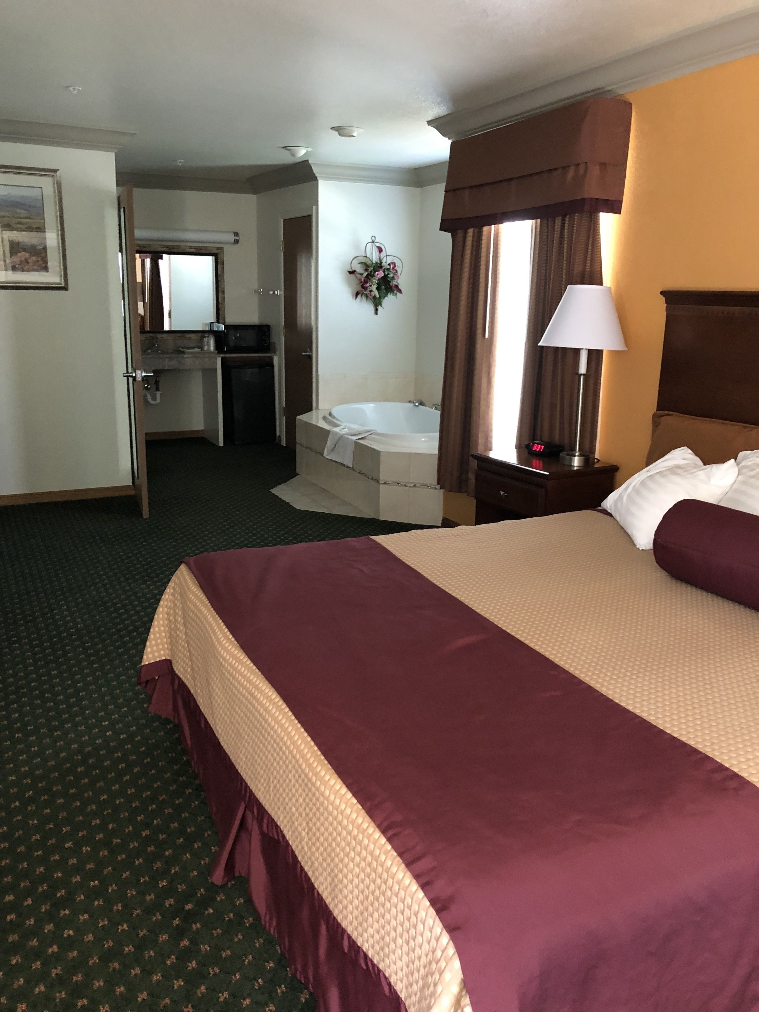 A hotel room with a large bed, a desk with a chair, a bathroom with a sink and mirror, and a window with curtains.