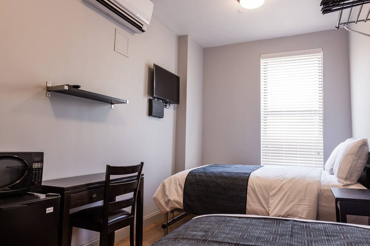 Een slaapkamer met een bed, een bureau met een magnetron, een stoel, een televisie en een raam met jaloezieën.