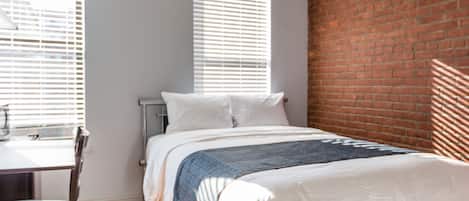 A bedroom with a bed, a desk, and a chair. There are two windows with blinds.