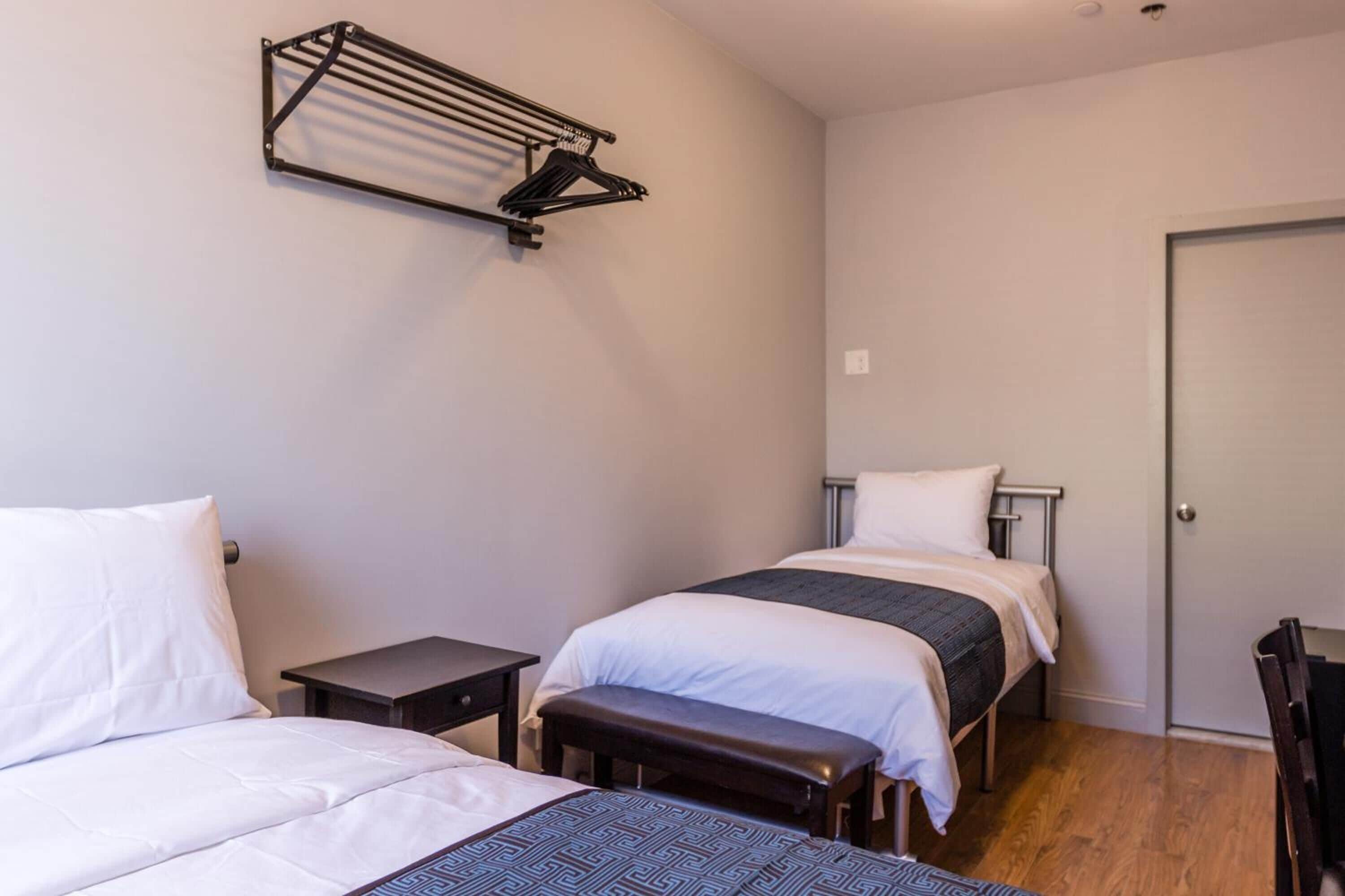 Ein Hotelzimmer mit zwei Betten, einer Bank, einem kleinen Tisch und einem an der Wand befestigten Wäscheständer.