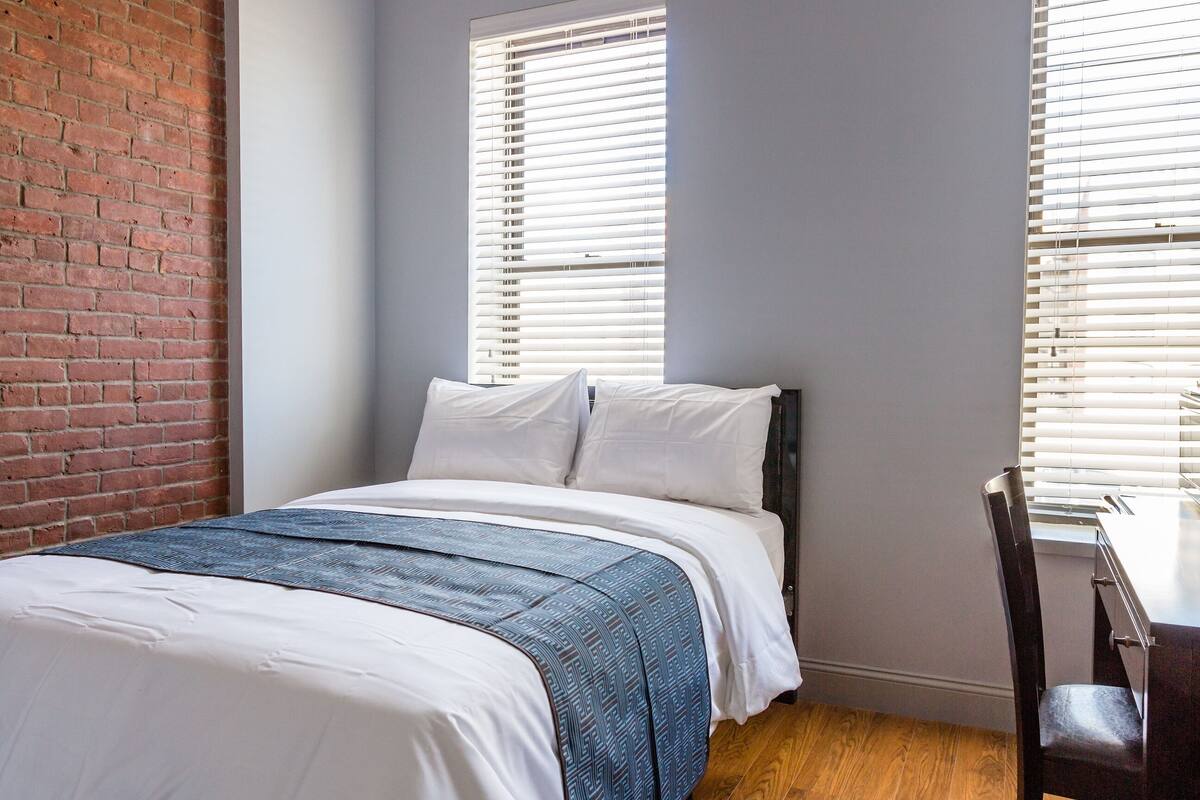 Een slaapkamer met een bakstenen muur, twee ramen met zonwering, een bed met wit beddengoed, een blauwe geborduurde plaid en een houten vloer.