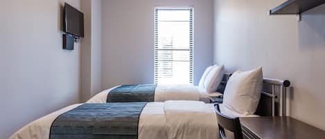 A small, compact room with two single beds, a TV mounted on the wall, and a window with blinds.