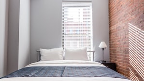 Un dormitorio con cama, ropa de cama blanca, una ventana con persianas, una mesita de noche con lámpara y una pared de ladrillo.