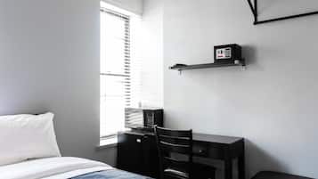 Ein Schlafzimmer mit einem Bett, einem Schreibtisch, einem Stuhl, einem Regal mit einem Gerät und einem Fenster mit Jalousien.