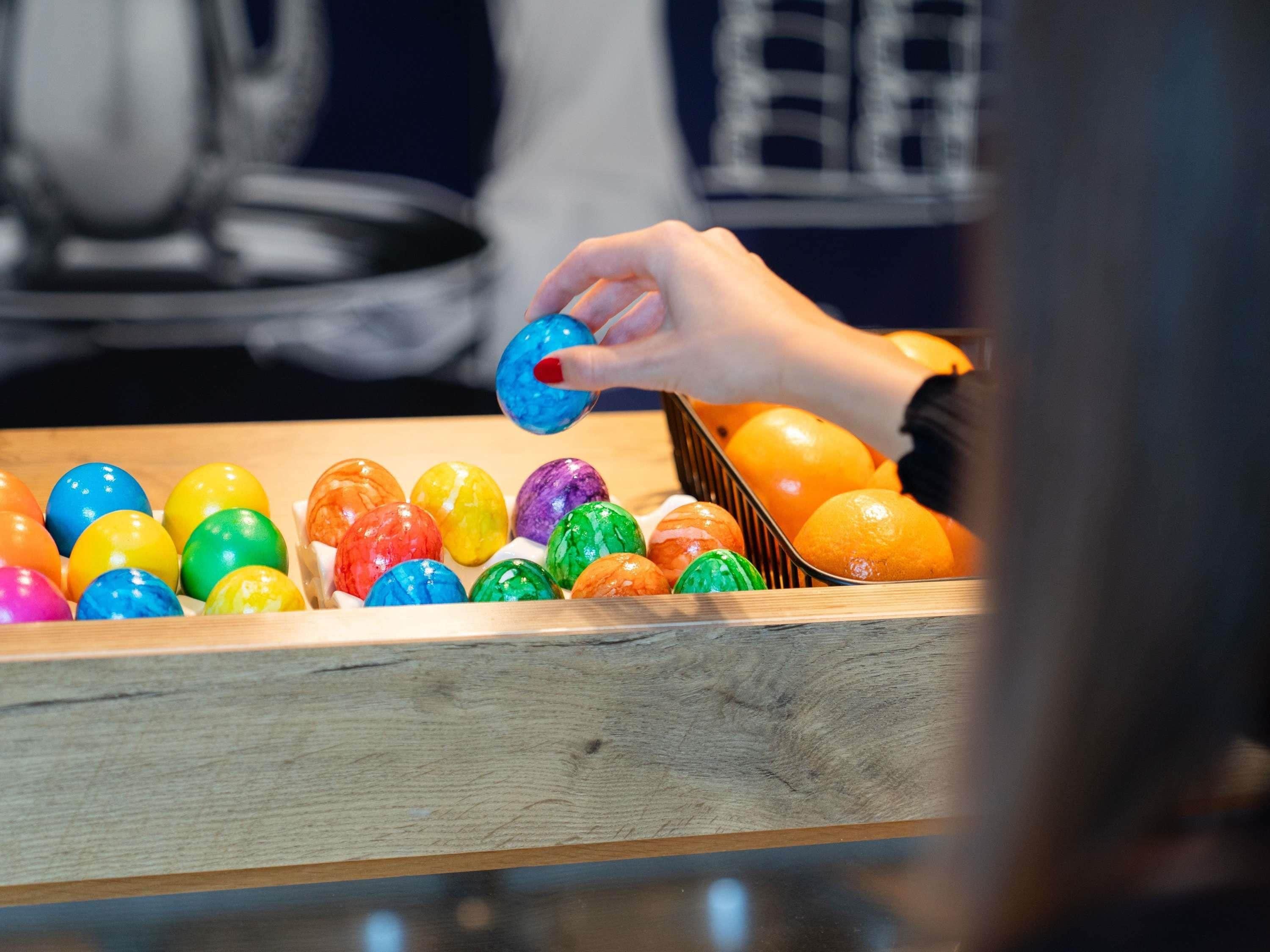 Una mano selecciona un huevo colorido de una caja de madera llena de huevos de diversos colores.