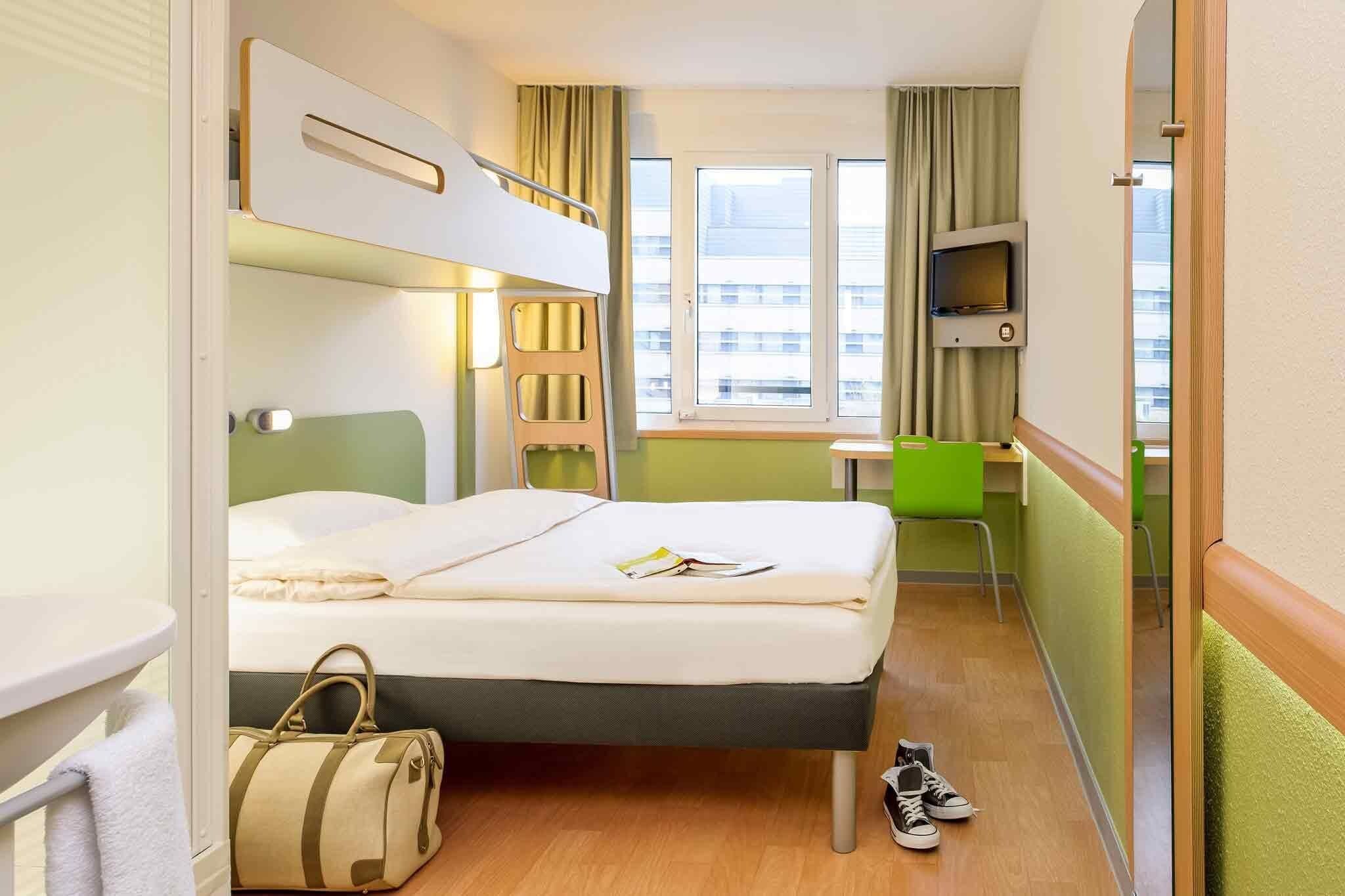A hotel room with a bunk bed, a desk, a chair, a TV, and a window with curtains.
