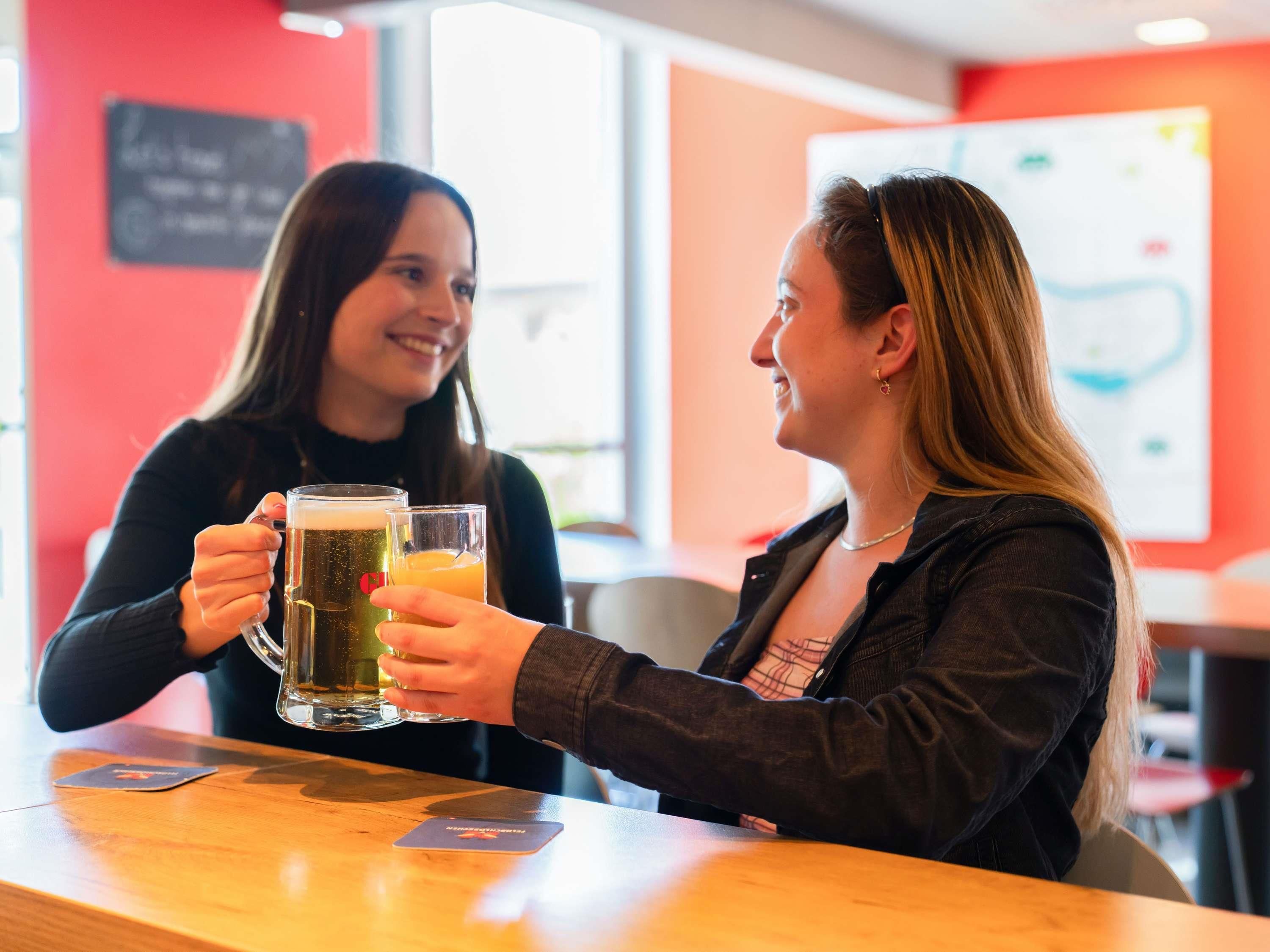 Two people toasting with beer mugs at a bar with a red wall and a whiteboard in the background.