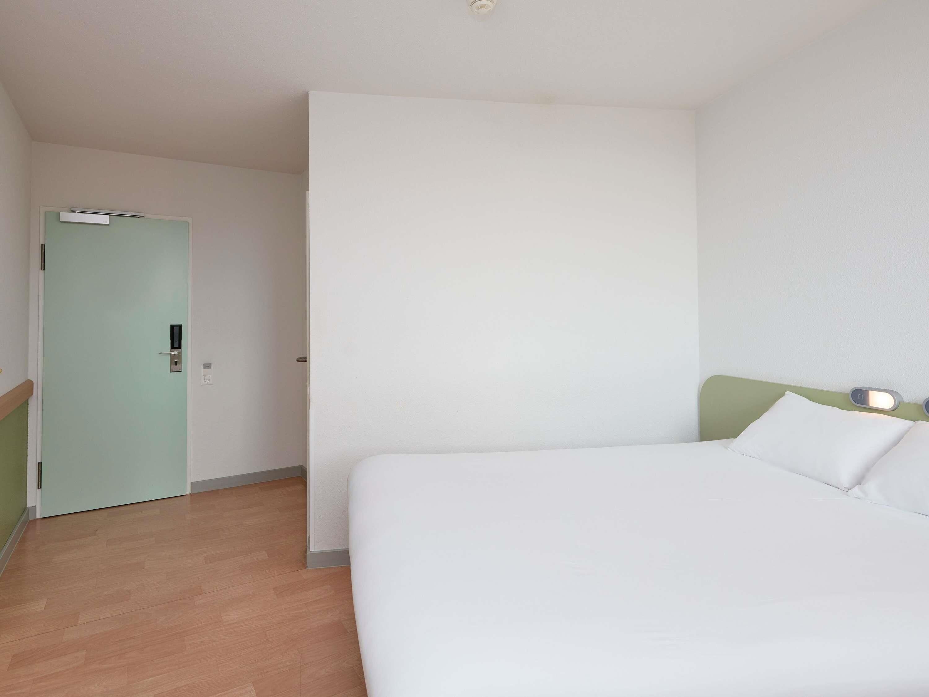 A hotel room with a bed, a door, and a light switch on the wall.