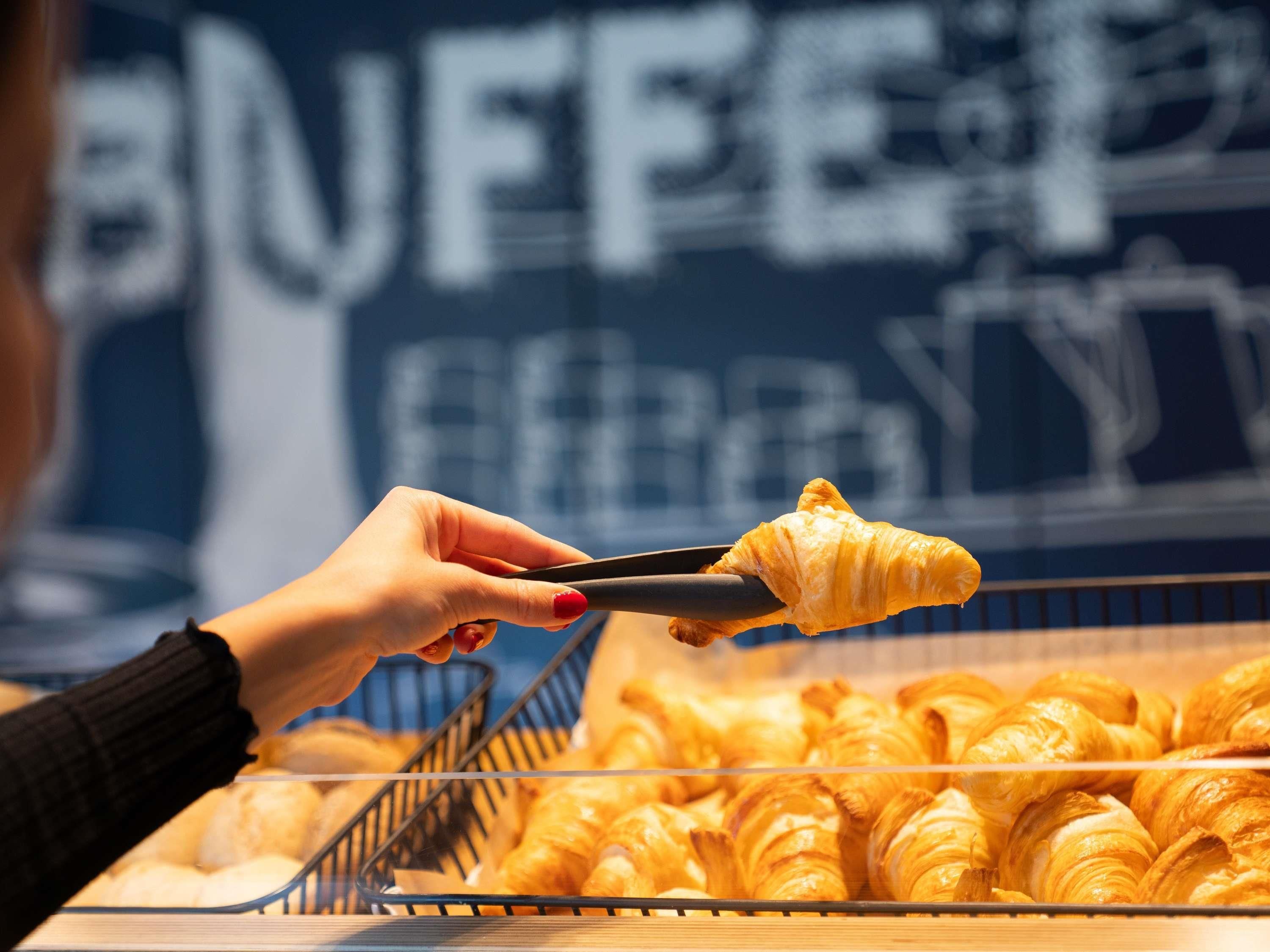 Una persona sosteniendo un croissant con unas pinzas sobre una bandeja de croissants.
