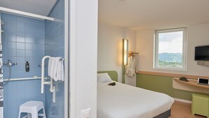 A hotel room with a bed, a window with a view, a bathroom with blue tiles, and a small stool. - ibis budget Bern Expo (Bern)