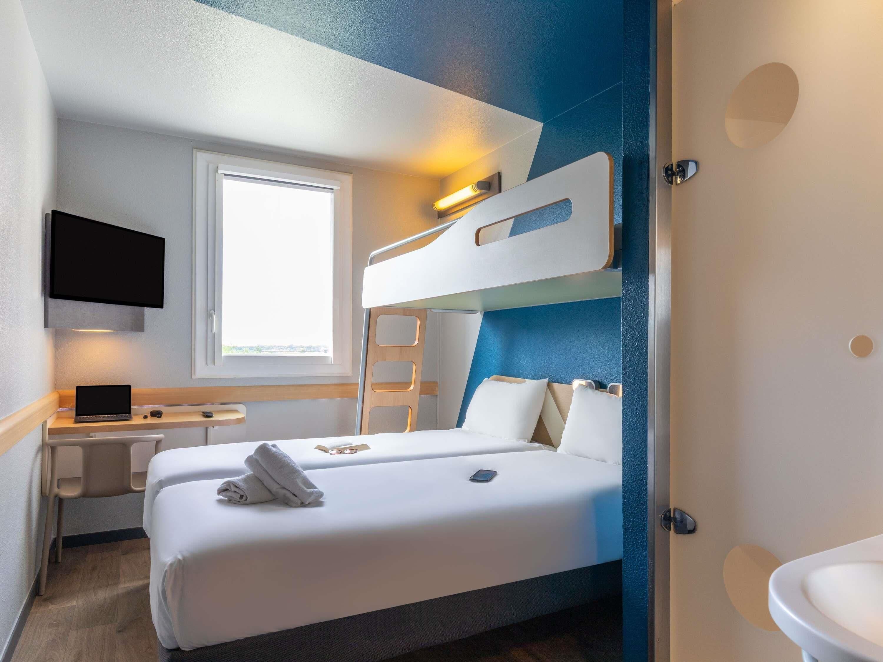 A hotel room with a bunk bed, a desk with a laptop, a window, and a bathroom.