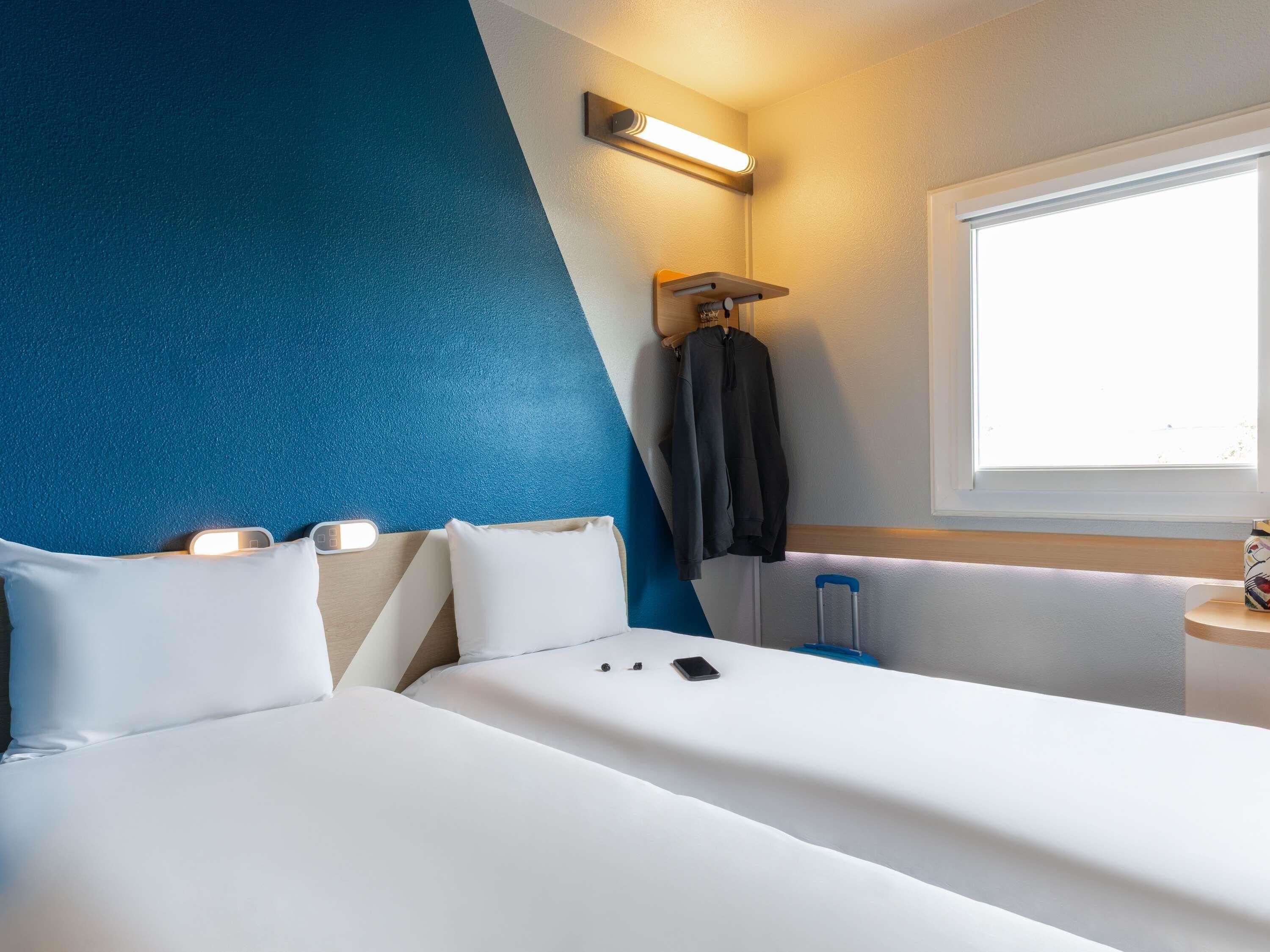 Habitació d'hotel amb un llit, almoines, una taula de nit, una làmpada, una finestra i una paret amb un detall blau.
