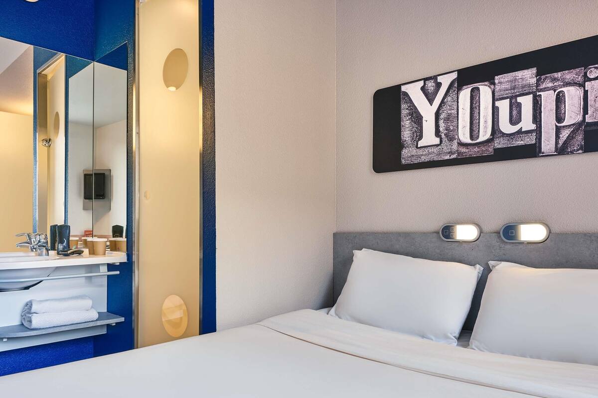 A hotel room with a bed, a bedside table, a mirror, and a wall-mounted sign.