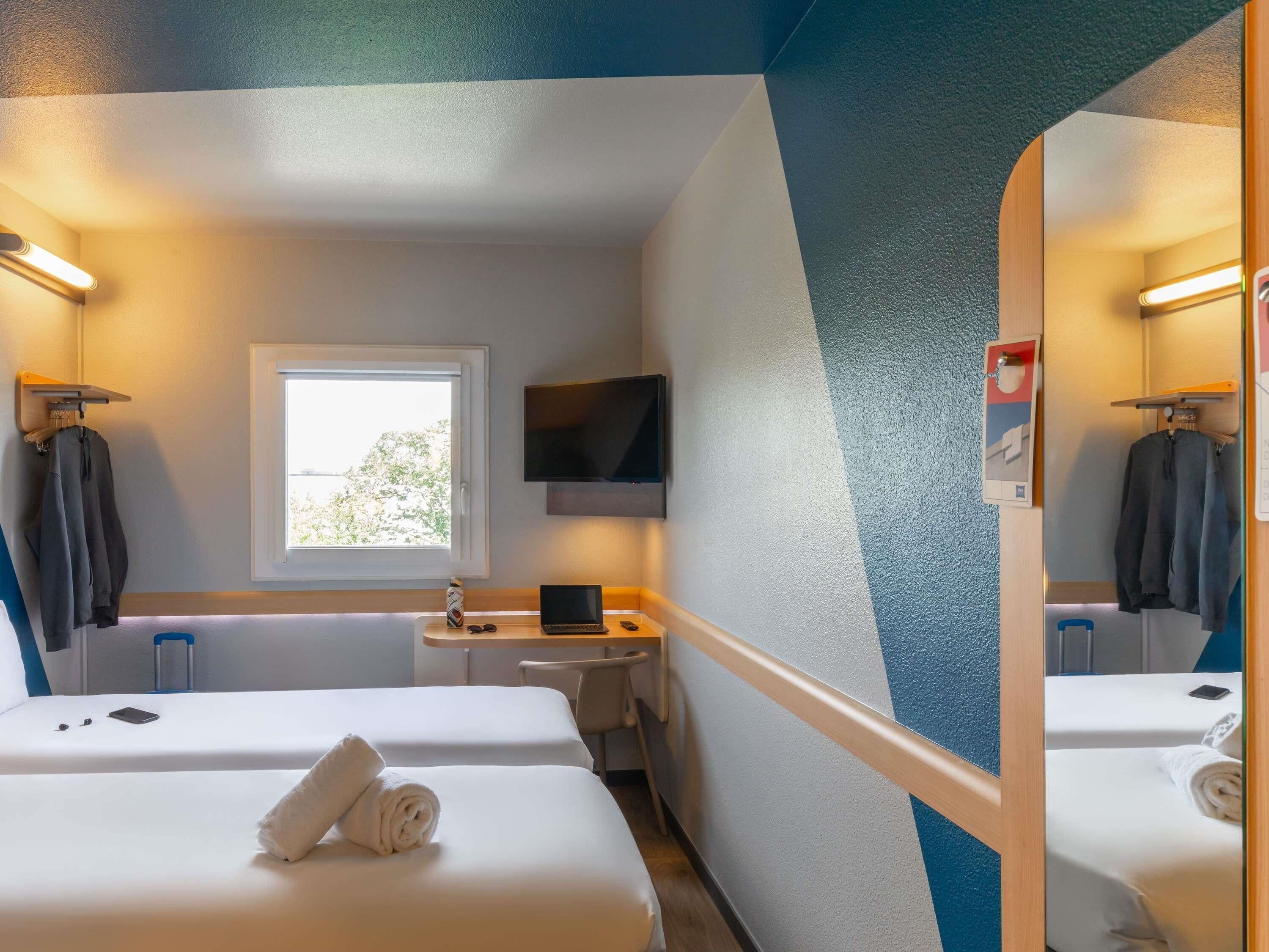 A hotel room with two beds, a desk, a TV, and a window.
