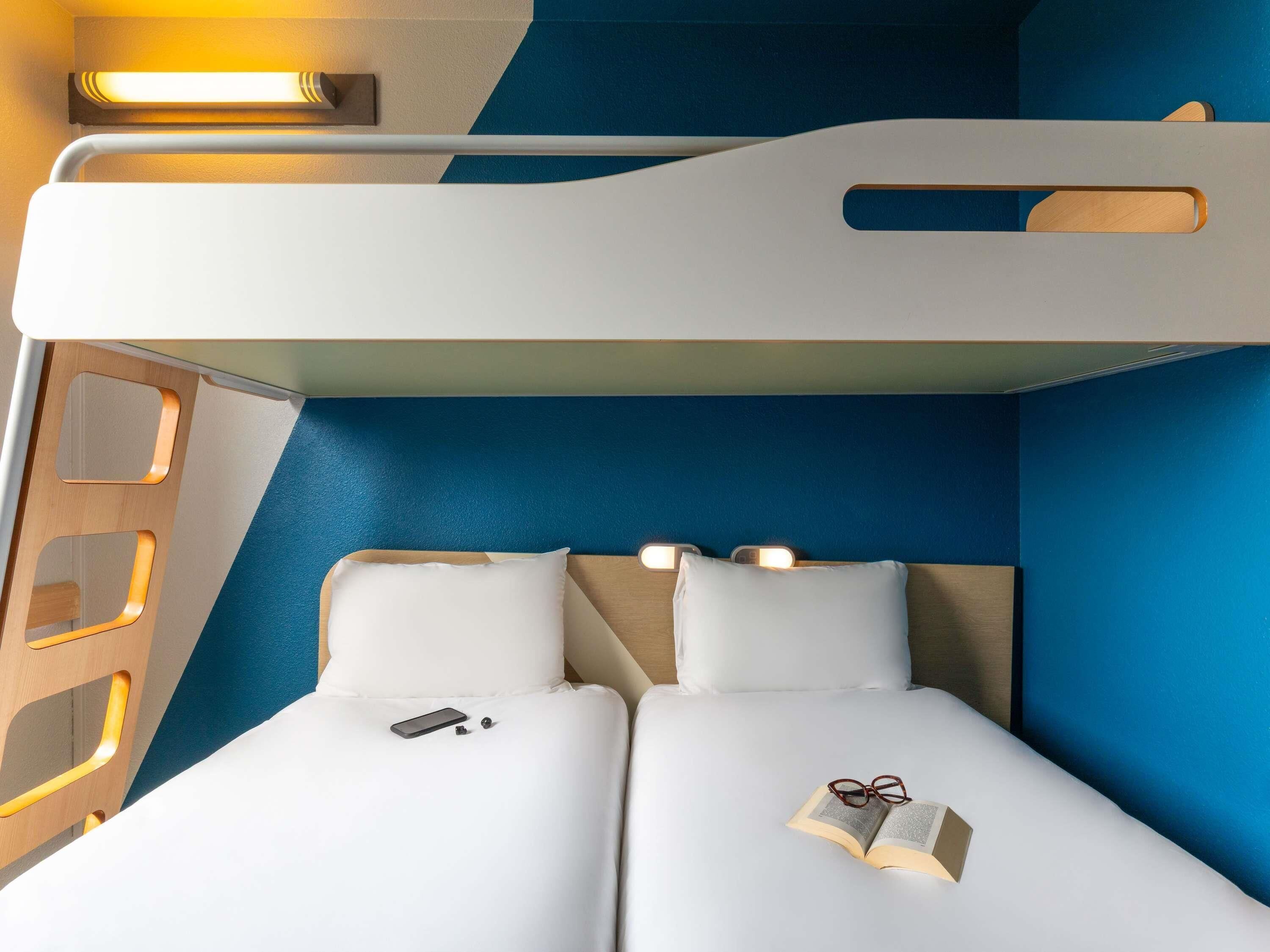 A bunk bed with a headboard, two pillows, a bedside table with a book and glasses, and a wall-mounted light fixture.