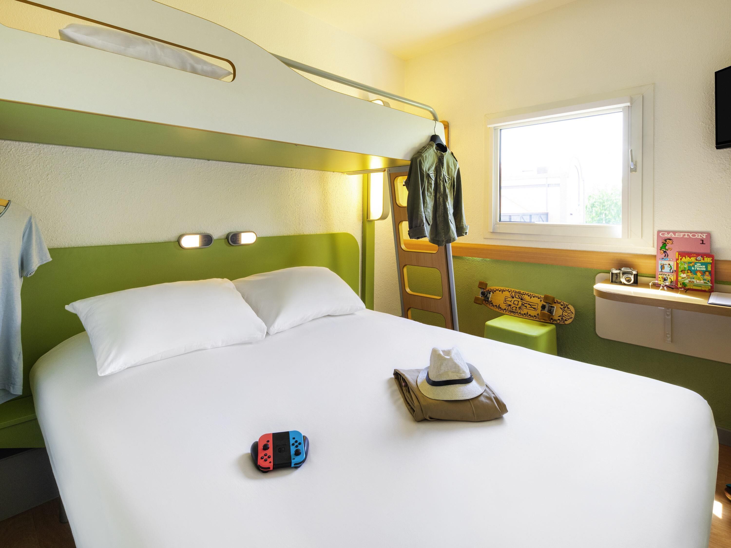 A neatly made bed with pillows, a folded hat, and a Nintendo Switch controller on the bed. There is a ladder against the wall, a window with daylight coming through, and a shelf with a camera and books.