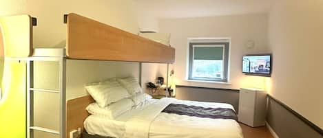 A compact hotel room with a bunk bed, a desk with a computer, a small refrigerator, and a window with a view.