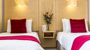 Habitación de hotel con dos camas, cada una con ropa de cama blanca y almohadas rojas. Hay una mesita de noche pequeña con un florero y una caja de pañuelos.