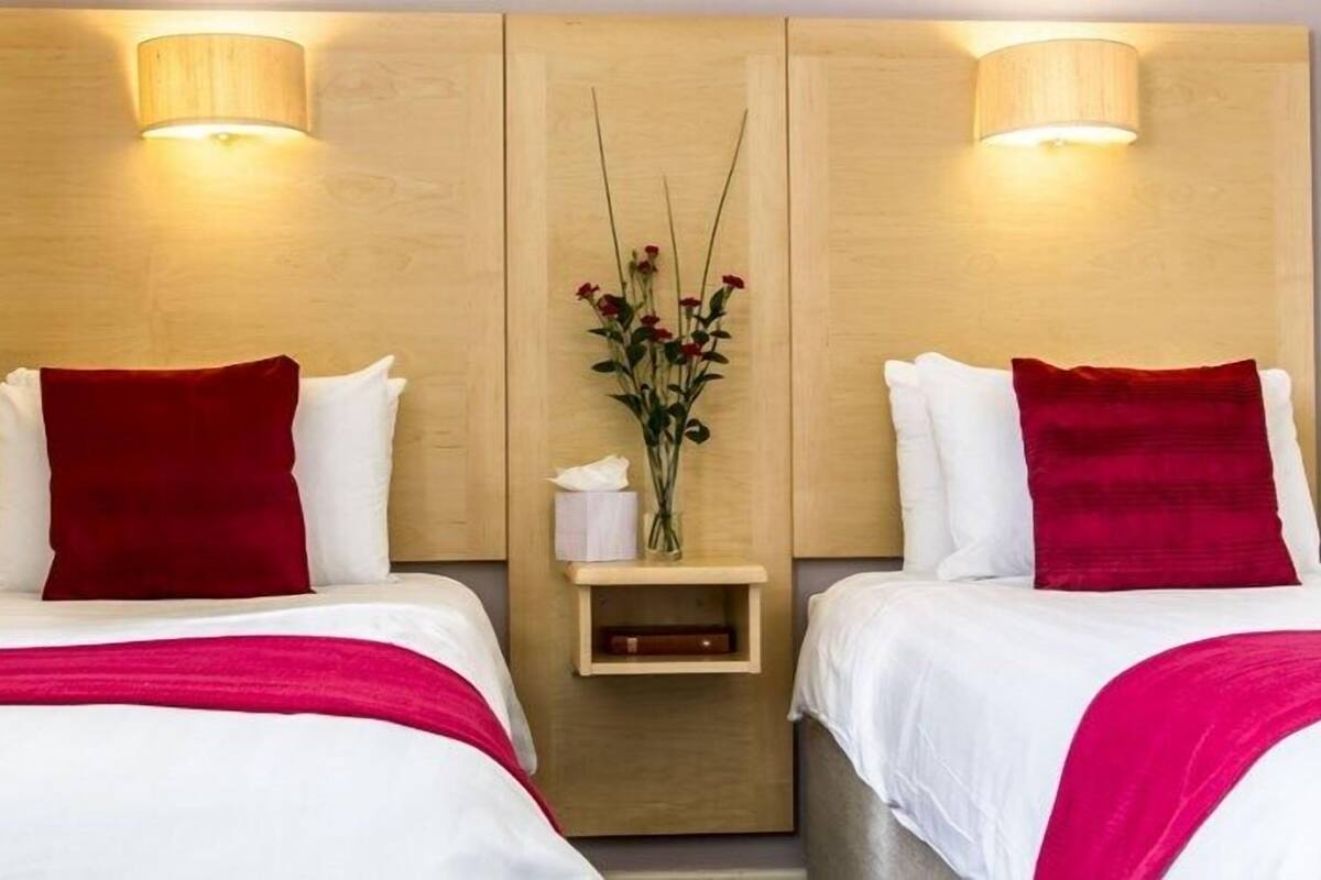 Habitación de hotel con dos camas, cada una con ropa de cama blanca y almohadas rojas. Hay una mesita de noche pequeña con un florero y una caja de pañuelos.