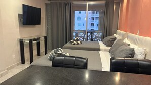 Moderni hotellihuone, jossa on sänky, sohva, televisio ja parveke, jolta on näkymä rakennuksiin.