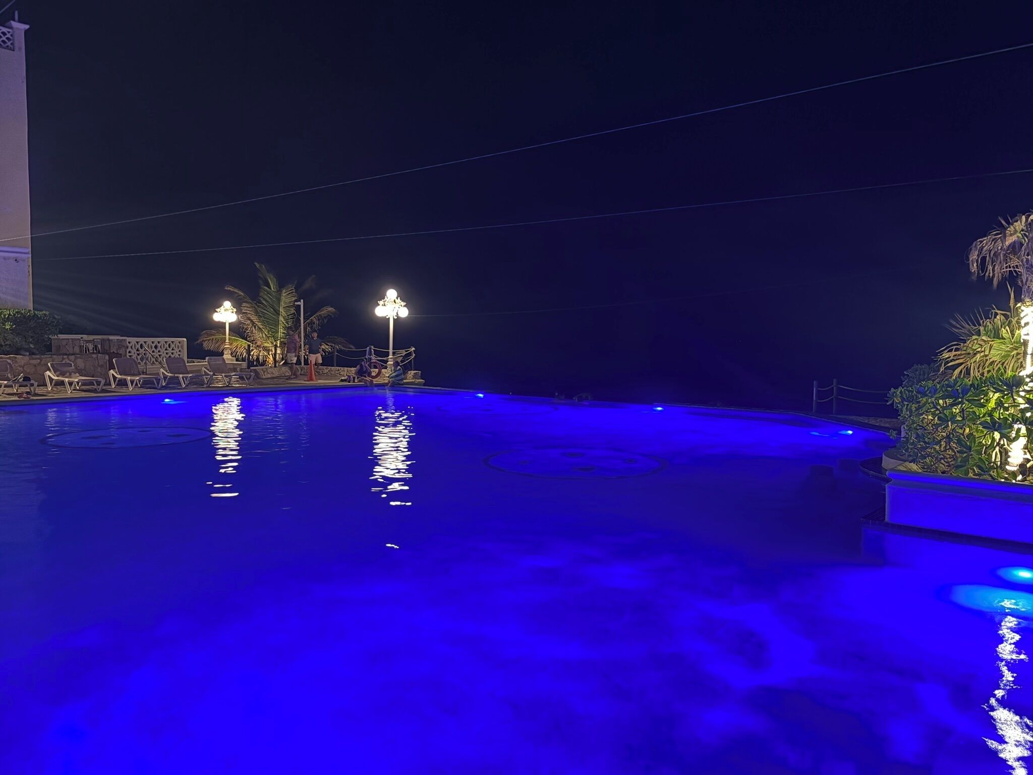 Una piscina illuminata di blu di notte, con sedie a sdraio e un lampione.