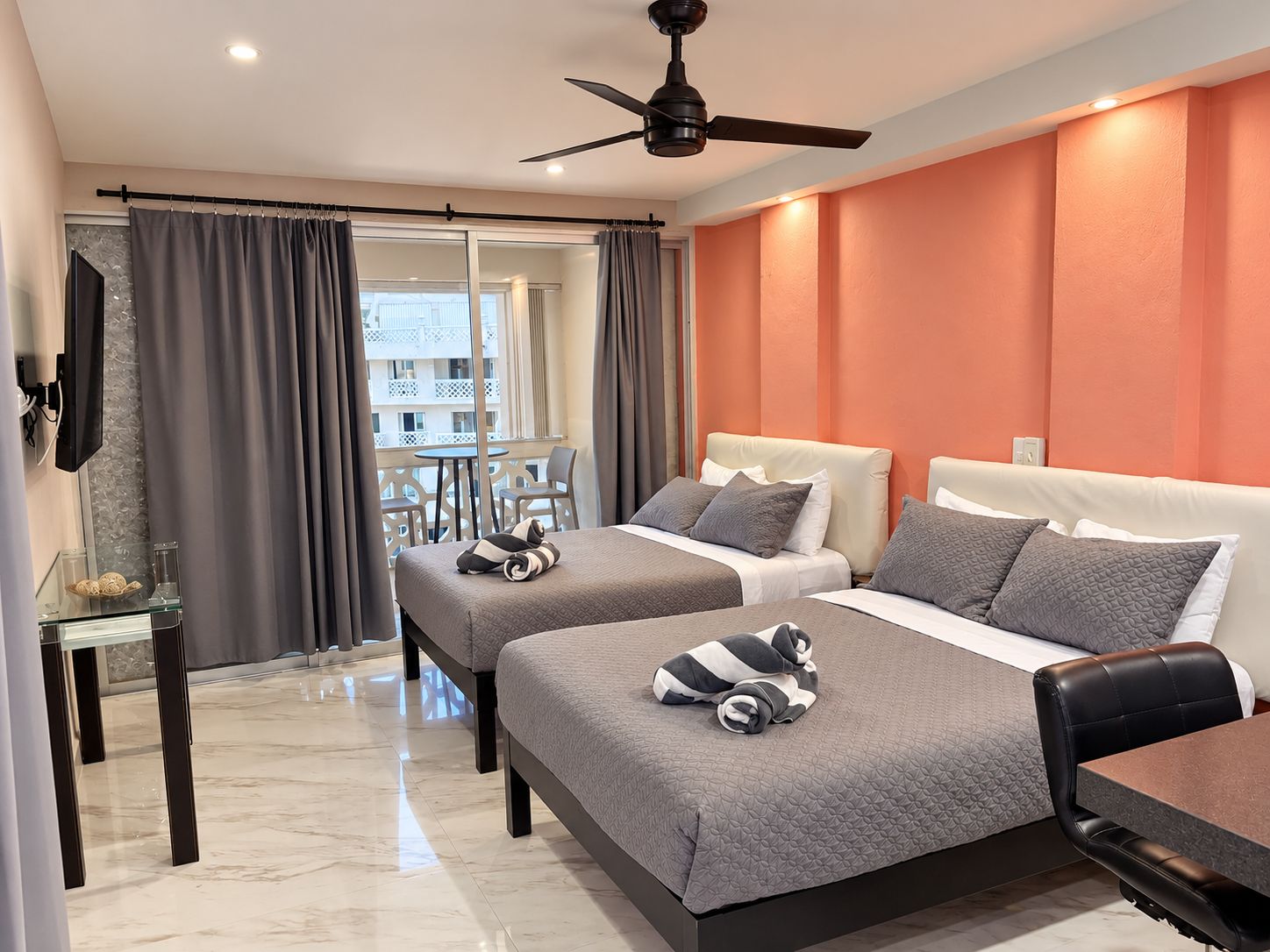 Une chambre d’hôtel avec deux lits, un ventilateur de plafond, une table en verre avec une chaise, et une vue sur le balcon extérieur.