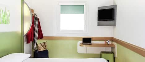 Quarto de hotel com uma cama, uma TV fixada na parede, uma mesinha com um laptop e uma janela com cortina.