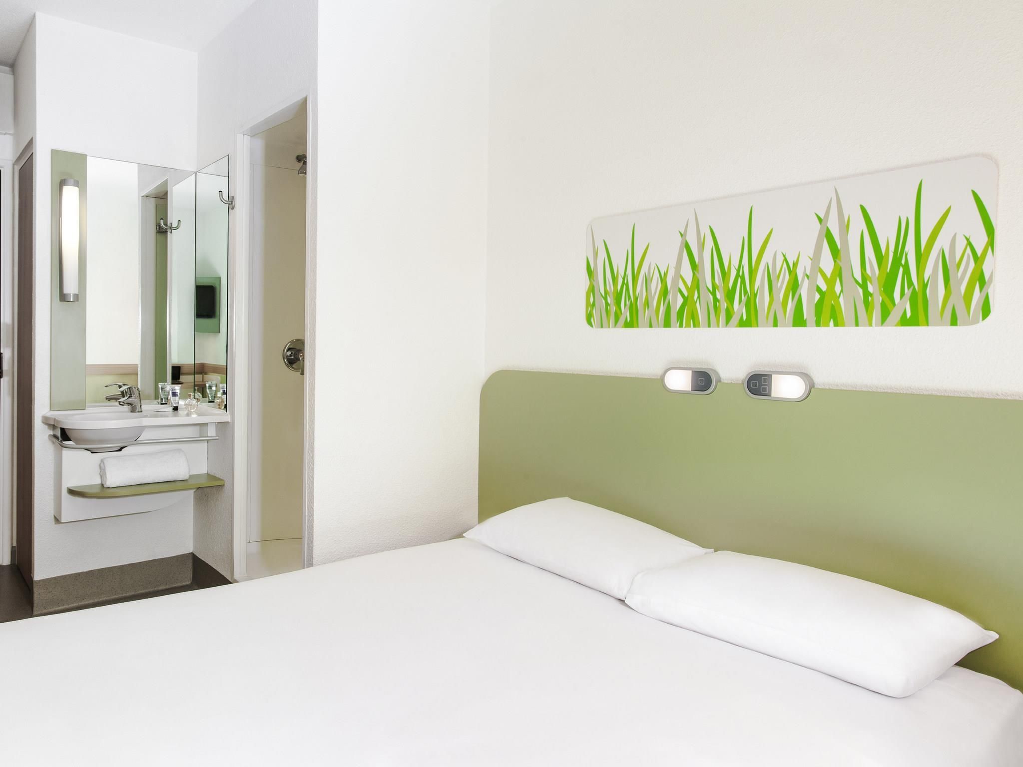 A hotel room with a bed, a bedside table, a mirror, and a wall-mounted light fixture.